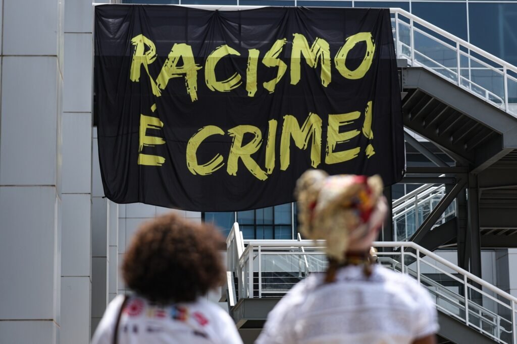 igualdade racial: Duas pessoas observam faixa preta com a frase "Racismo é crime!" em evento em Brasília.