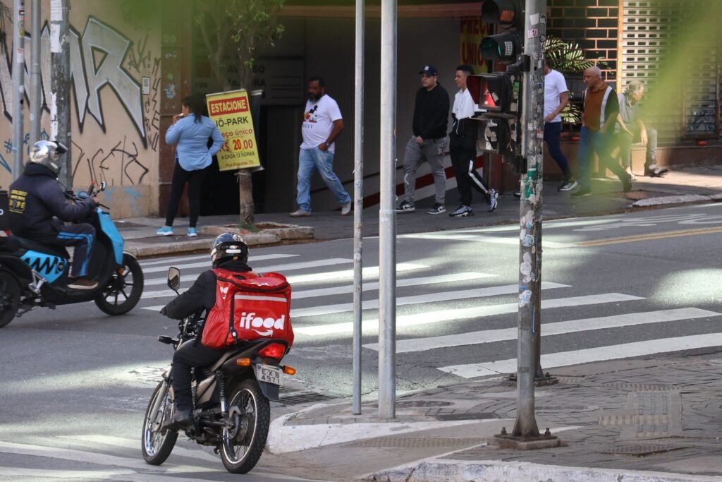 Motociclista com mochila vermelha da "ifood" em via urbana, com pedestres e outro motociclista ao fundo em um cruzamento.