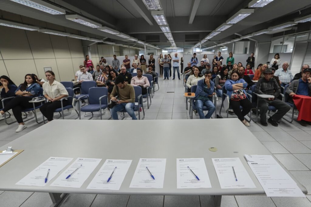 Bolsa Familia: Documentos com o logotipo da Caixa Econômica Federal e canetas sobre uma mesa, com pessoas aguardando ao fundo.