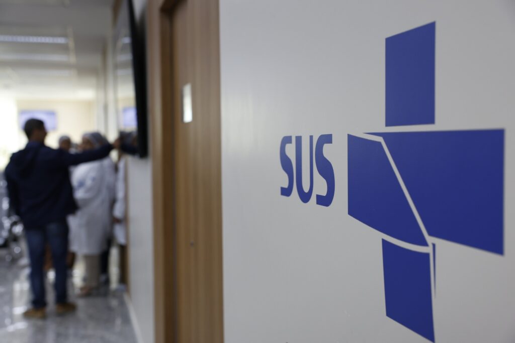 homens e o cancer de mama: Logotipo azul do SUS em parede branca, com pessoas desfocadas em um corredor ao fundo.