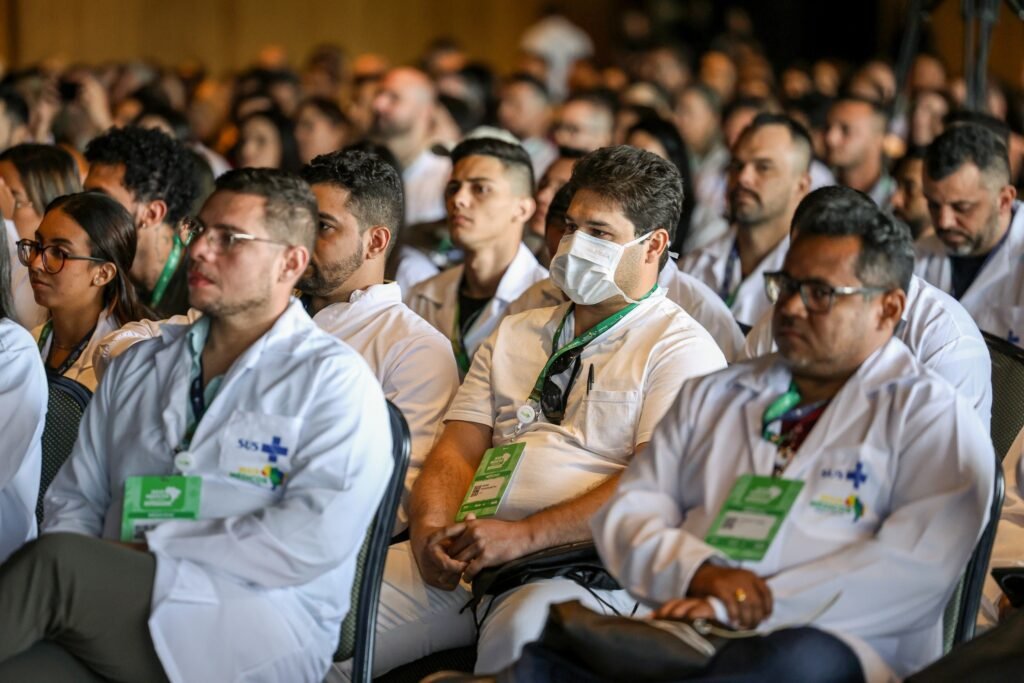 Mais Médicos Especialistas: profissionais de saúde com jalecos sentados em auditório durante anúncio do programa.