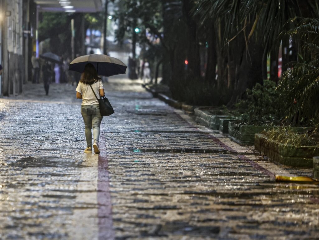 chuvas fortes: mulher de costas caminha sob um guarda-chuva preto em calçada de pedras molhada pela chuva.