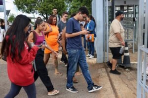 Enem 2026: jovens estudantes aguardam em fila ao ar livre, alguns consultando celulares.
