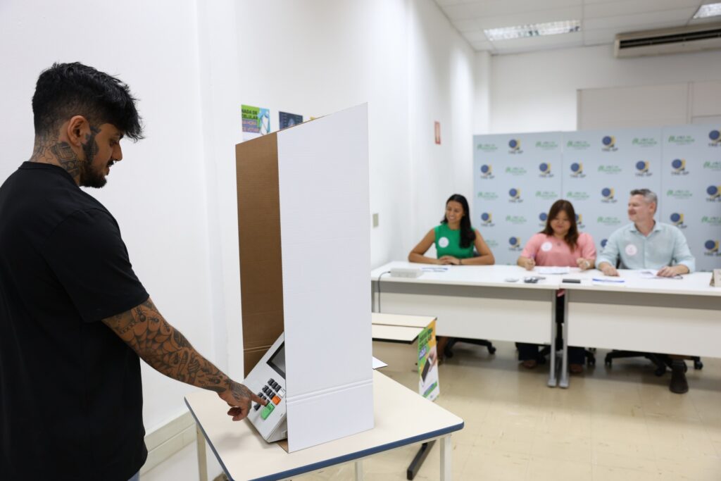 TRE-SP: homem com tatuagens vota em urna eletrônica em seção eleitoral cenográfica.