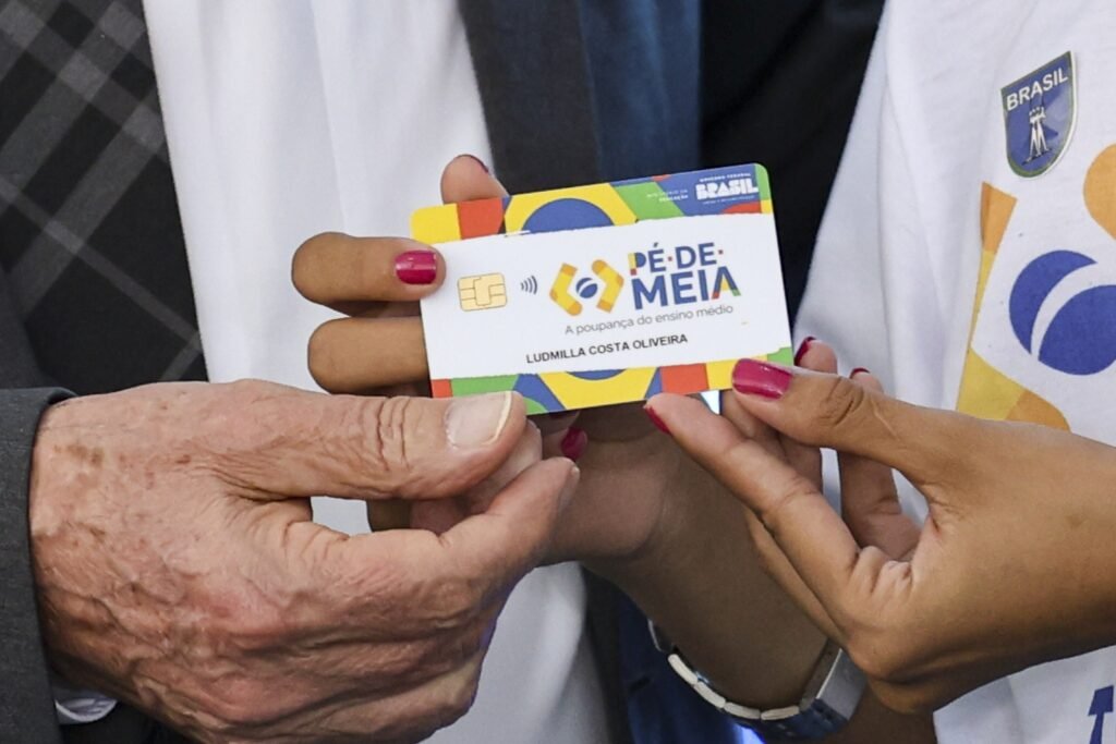 Close-up de mãos entregando um cartão do programa "Pé-de-Meia" com a frase "A poupança do ensino médio".