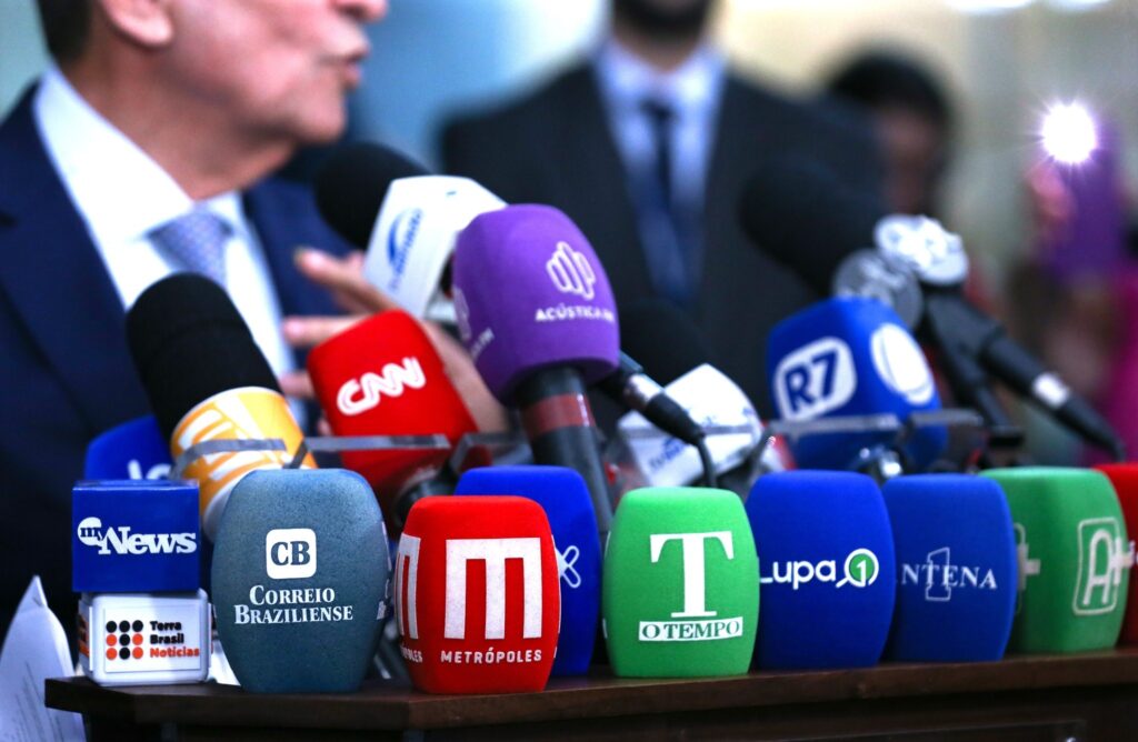 Democracia: Diversos microfones de veículos de imprensa, como "CNN", "R7" e "Metrópoles", posicionados à frente de um homem de terno que fala em uma coletiva de imprensa.