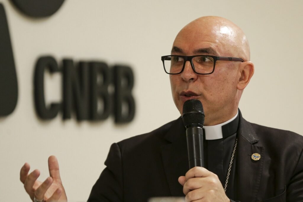 Dom Ricardo Hoepers, com óculos e traje clerical, fala ao microfone diante de um fundo com a sigla 'CNBB'.