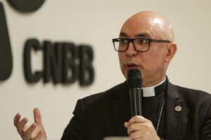 Dom Ricardo Hoepers, com óculos e traje clerical, fala ao microfone diante de um fundo com a sigla 'CNBB'.