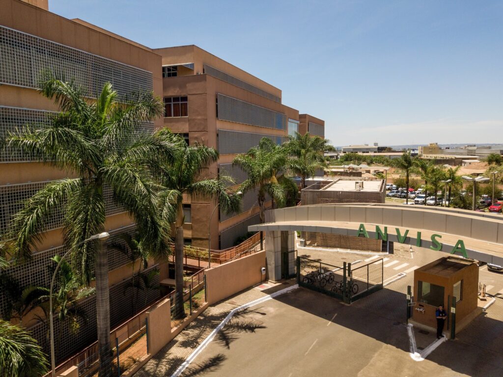 Fachada do prédio da Anvisa com letreiro verde "ANVISA" na entrada, cercado por palmeiras.