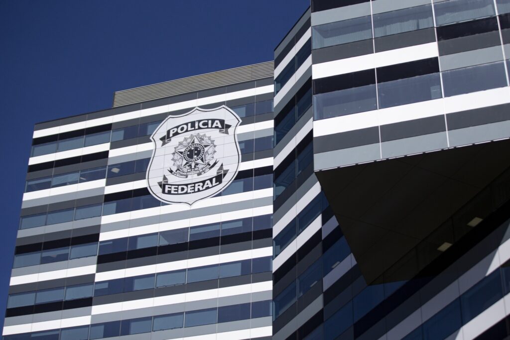 Fachada de prédio moderno com faixas horizontais brancas e escuras, exibindo o logotipo "POLICIA FEDERAL" com o brasão da República.