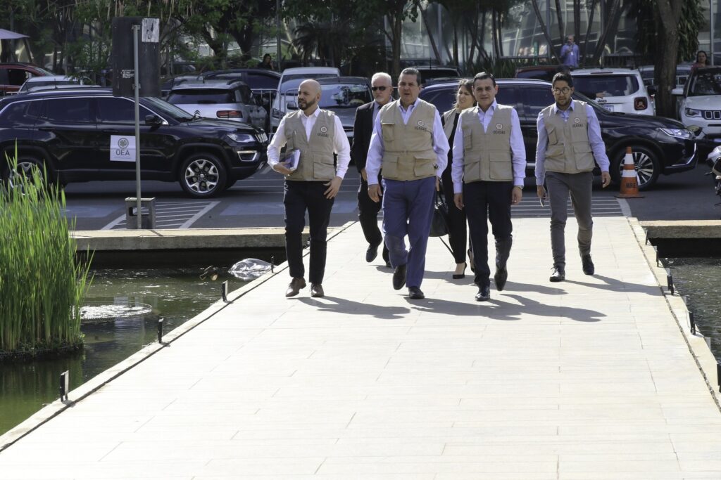 denúncia contra a Meta: Grupo de pessoas, algumas vestindo coletes com a sigla OEA OAS, caminha por passarela de pedra.
