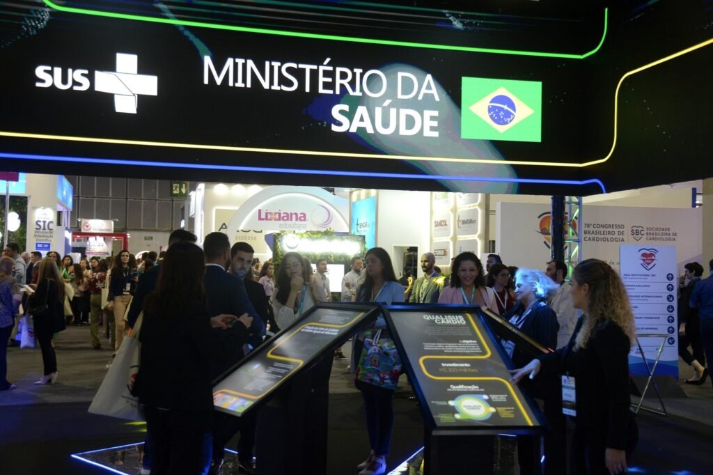 doencas cardiovasculares: Pessoas em um evento de medicina observam telas digitais sob um banner escrito "MINISTÉRIO DA SAÚDE".