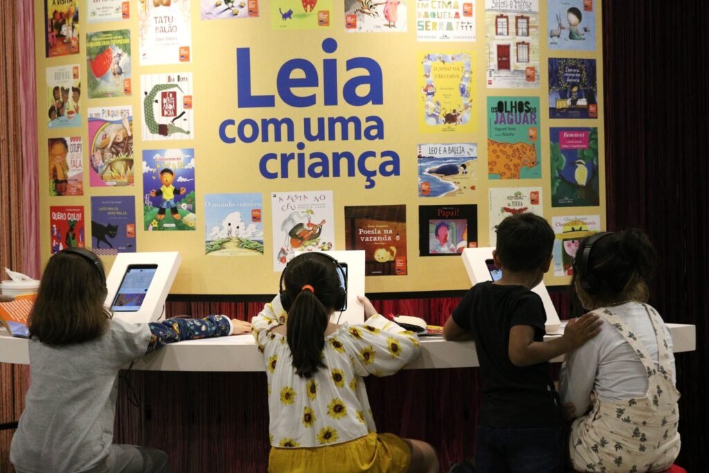 educação infantil: crianças de costas usam tablets diante de painel com capas de livros infantis.