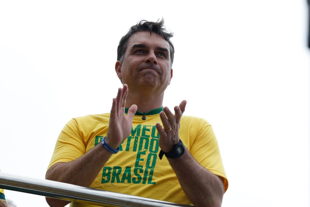 Flávio Bolsonaro veste camiseta amarela com os dizeres "Meu partido é o Brasil" e aplaude em evento político.