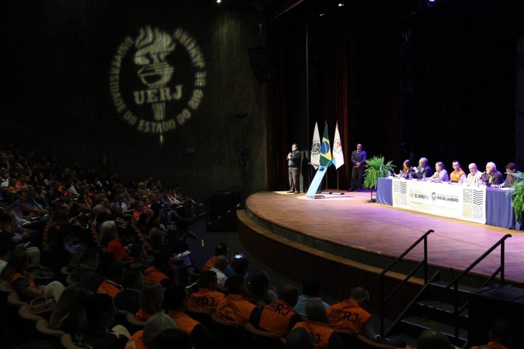 emergencia: Reunião do comitê de Defesa Civil sobre efeitos climáticos em auditório com público e autoridades.