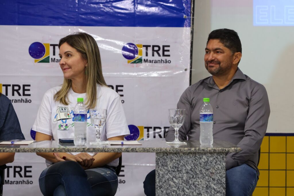 assédio eleitoral: Uma mulher e um homem sentados à mesa em frente a um banner institucional da Justiça Eleitoral.