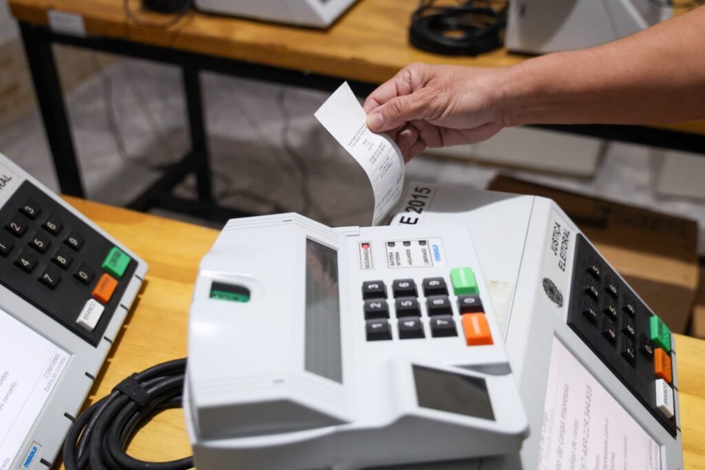 reforço de segurança nas eleições: urna eletrônica com comprovante de papel e inscrição da Justiça Eleitoral sobre mesa de madeira.
