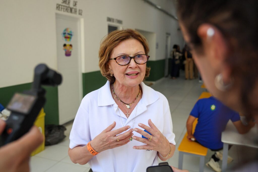 TRE-BA: Mulher de óculos e camisa polo branca gesticula em corredor, usando pulseira laranja com a sigla TRE.