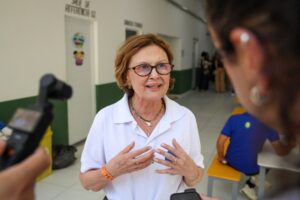 TRE-BA: Mulher de óculos e camisa polo branca gesticula em corredor, usando pulseira laranja com a sigla TRE.