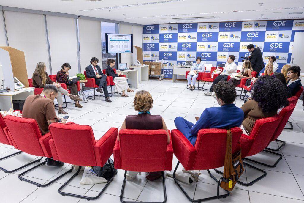 TSE: Pessoas sentadas em círculo em poltronas vermelhas durante evento institucional com painel ao fundo.