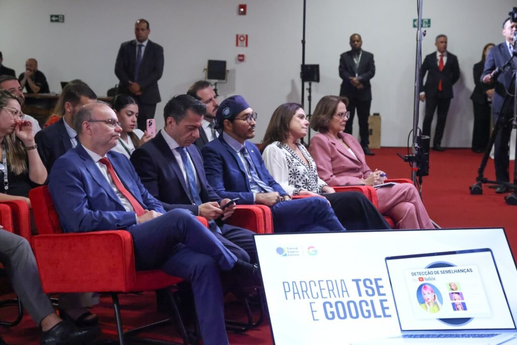 Pessoas em traje formal sentadas em poltronas vermelhas durante evento com tela exibindo "PARCERIA TSE E GOOGLE" e "DETECÇÃO DE SEMELHANÇAS no YouTube".