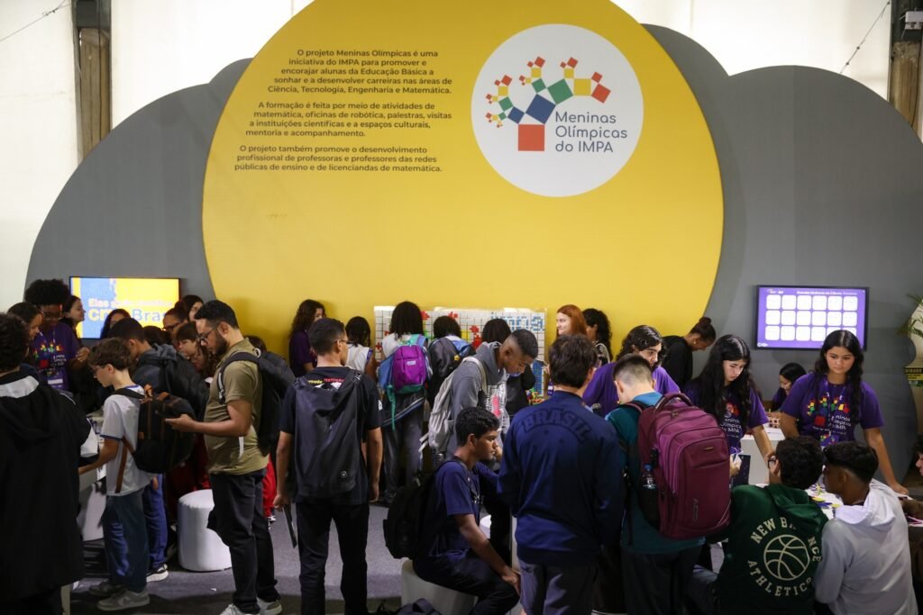 obmep: Jovens reunidos em estande do projeto Meninas Olímpicas do IMPA, com painel informativo amarelo ao fundo e telas digitais.