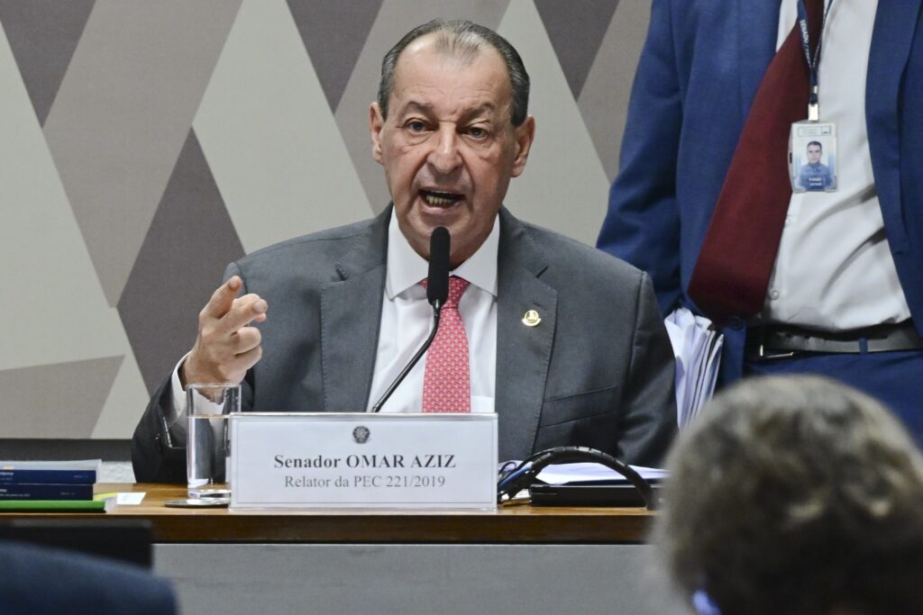 O senador Omar Aziz fala ao microfone durante reunião, com uma placa à sua frente com os dizeres "Senador OMAR AZIZ Relator da PEC 221/2019".