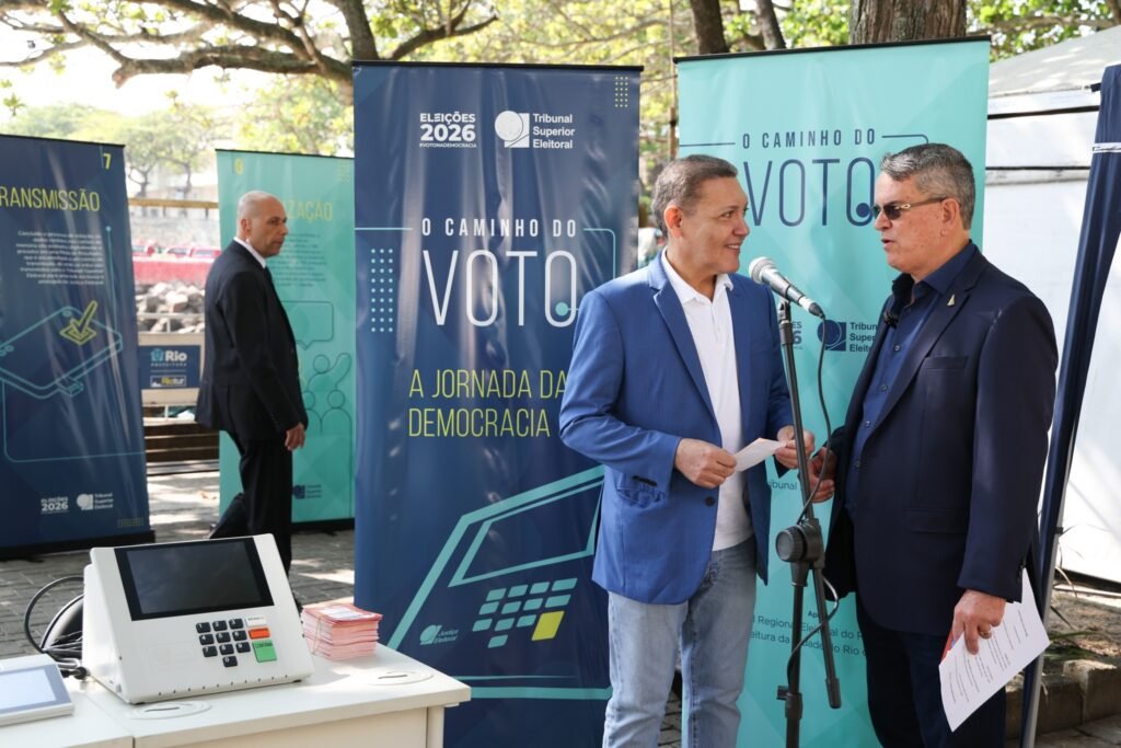 locais de votação: homens conversam ao lado de urna eletrônica e banners do Tribunal Superior Eleitoral em evento público.