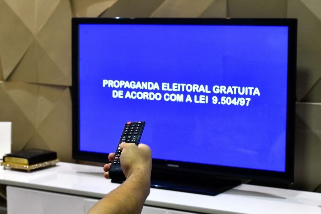 Mão segura controle remoto apontado para televisão com fundo azul e o texto "PROPAGANDA ELEITORAL GRATUITA DE ACORDO COM A LEI 9.504/97".