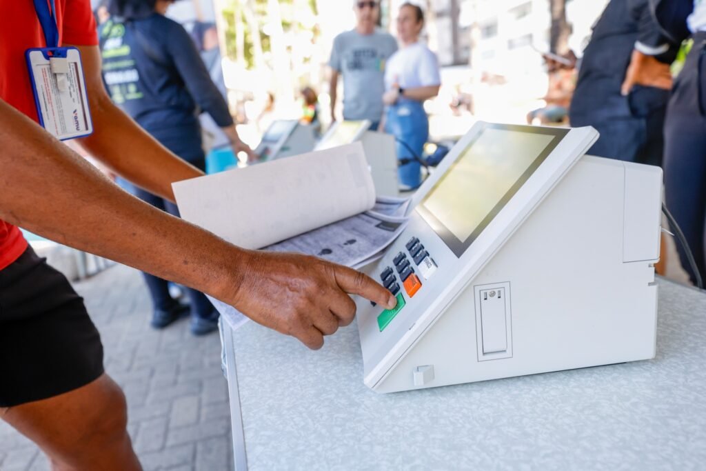 Pessoa com camiseta vermelha aperta a tecla verde de uma urna eletrônica em demonstração pública.