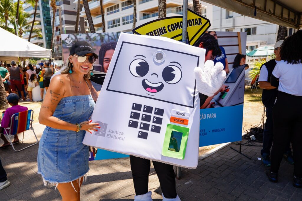 local de votacao: mulher sorri ao lado de mascote em formato de urna eletrônica com a inscrição Justiça Eleitoral.