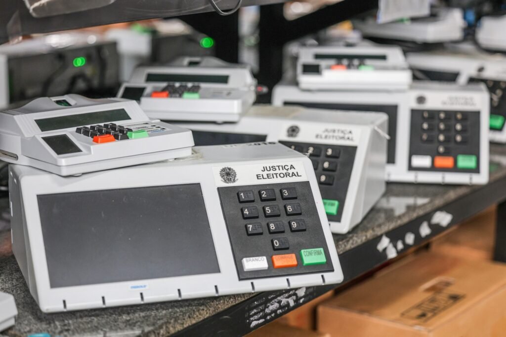 Diversas urnas eletrônicas da "JUSTIÇA ELEITORAL" alinhadas sobre uma bancada.