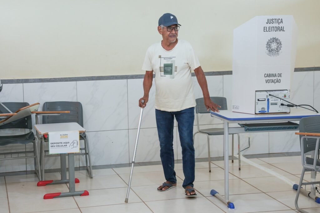 abstenção de idosos: Homem idoso com boné e muleta em seção eleitoral ao lado de urna e cabine de votação.
