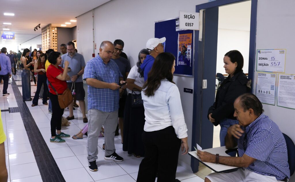 voto em trânsito: eleitores aguardam em fila em corredor de seção eleitoral diante de placas informativas.