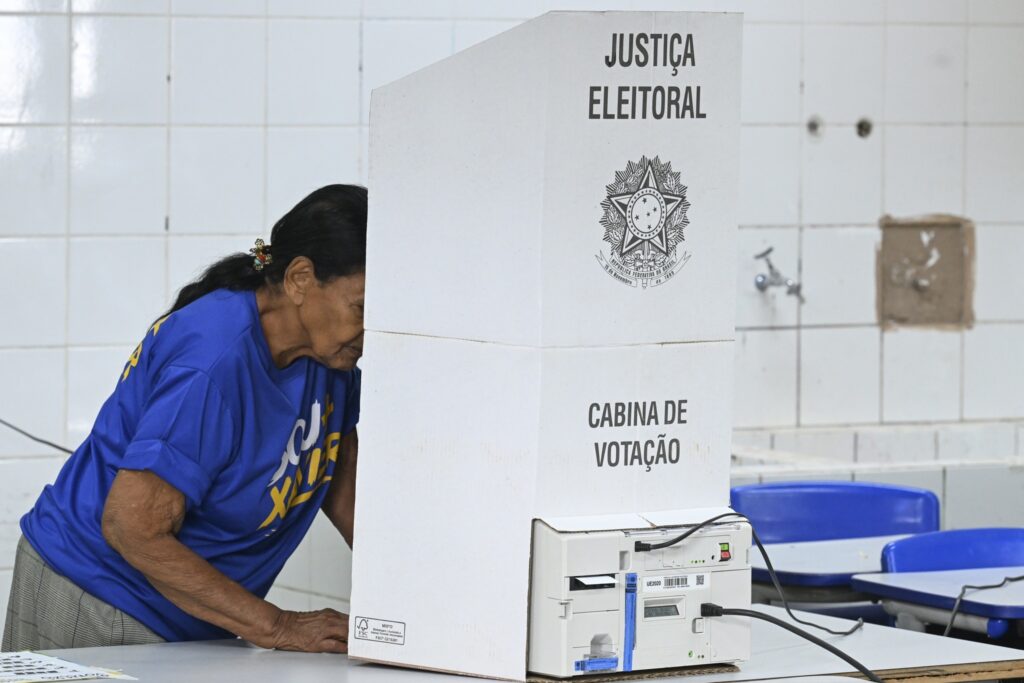 Mulher idosa vota em urna eletrônica dentro de uma cabine com os dizeres "JUSTIÇA ELEITORAL" e "CABINA DE VOTAÇÃO".