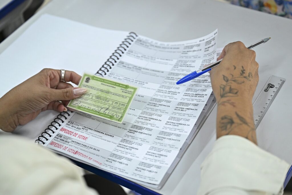 colinha para votar: pessoa segura título de eleitor verde e caneta sobre caderno com listas de nomes de eleitores em seção de votação.