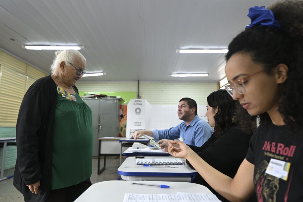 transporte em Salvador: eleitora aguarda diante de mesários em seção eleitoral com cabine de votação ao fundo.