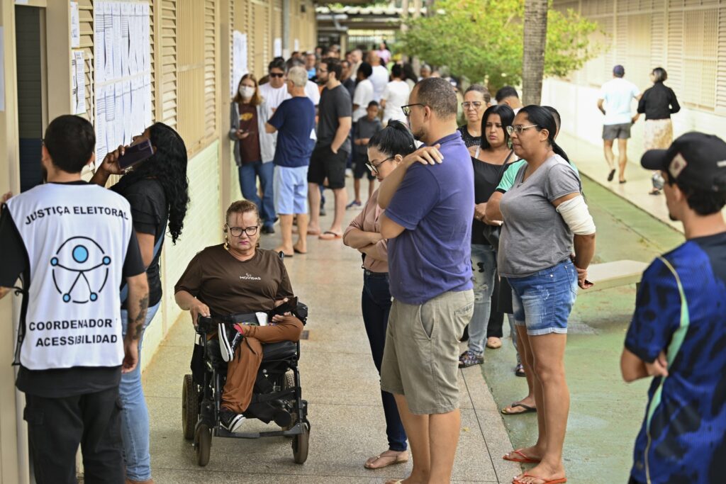 direitos de idosos: eleitores aguardam em fila para votação, com uma mulher em cadeira de rodas e um coordenador de acessibilidade.