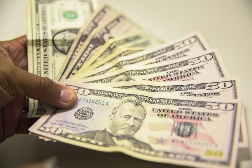 Mão segura um leque de notas de dólar americano, destacando cédulas de cinquenta dólares em primeiro plano.