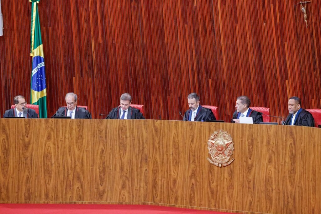 decisão do TSE: Seis ministros sentados à mesa semicircular em plenário com painel de madeira e a bandeira do Brasil.