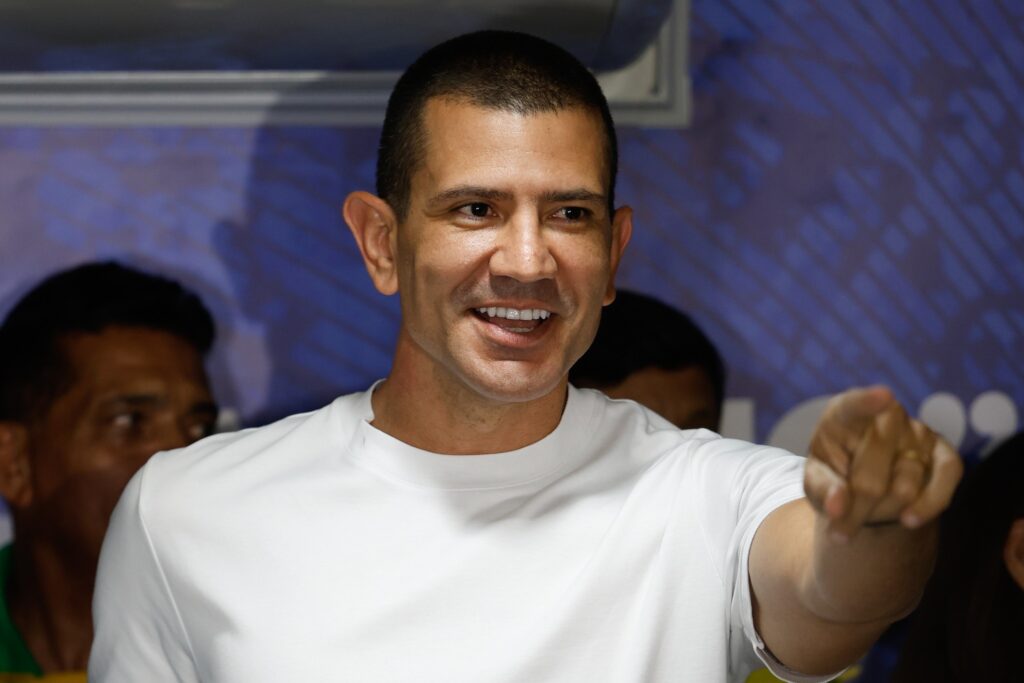 O candidato Douglas Ruas, de camisa branca, sorri e aponta para a frente em ambiente interno.
