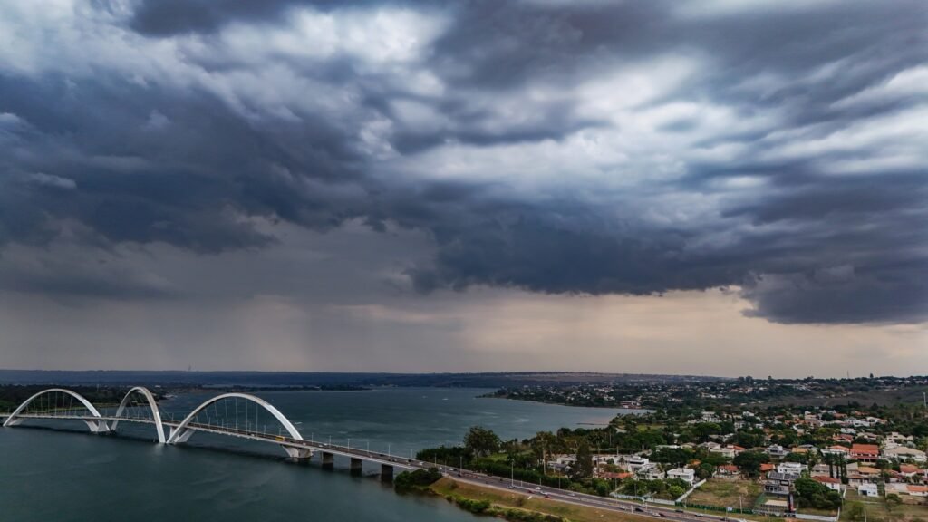 chuvas: Vista aérea da Ponte Juscelino Kubitschek sobre o Lago Paranoá, em Brasília, sob um céu coberto por nuvens escuras.