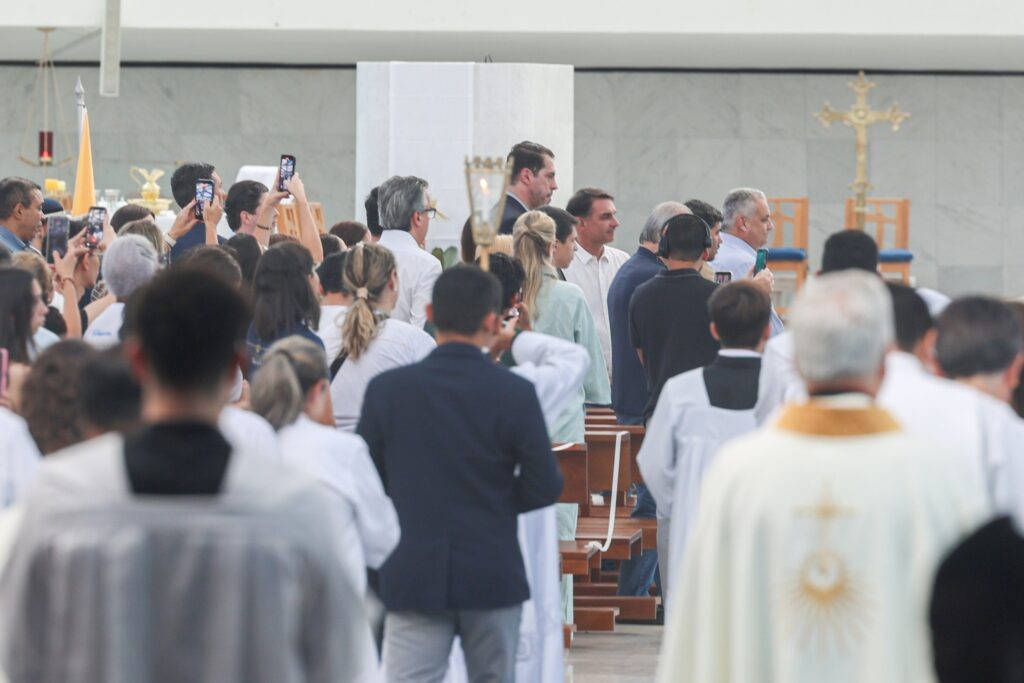 O senador Flávio Bolsonaro participa de missa na Catedral Metropolitana de Brasília, rodeado por fiéis.