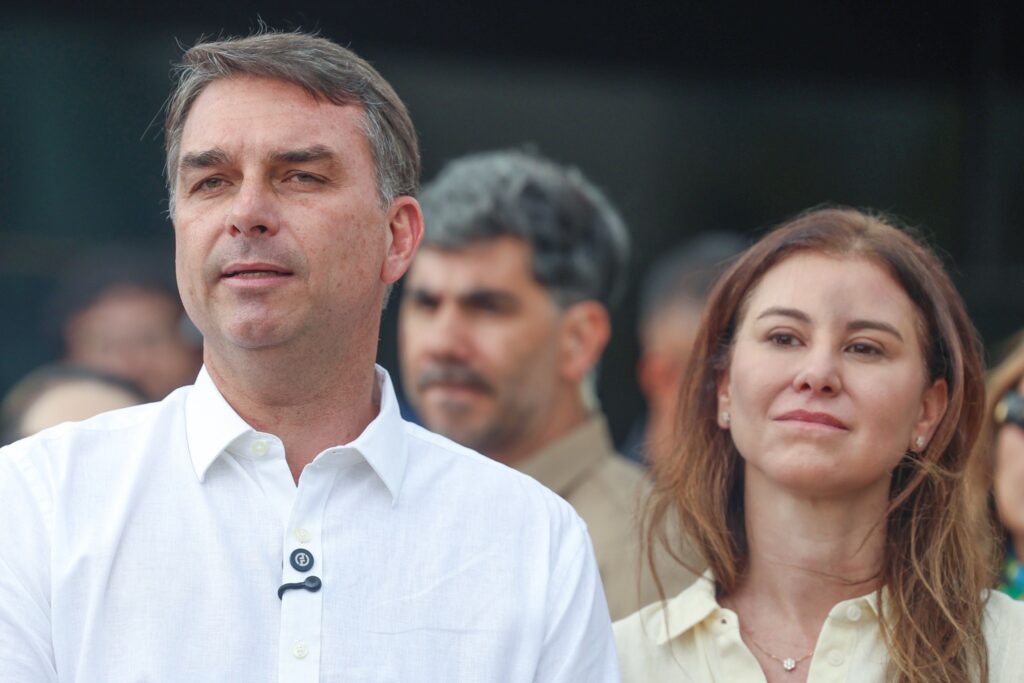 O senador Flávio Bolsonaro aparece em primeiro plano, vestindo camisa branca, ao lado de uma mulher em ambiente externo.