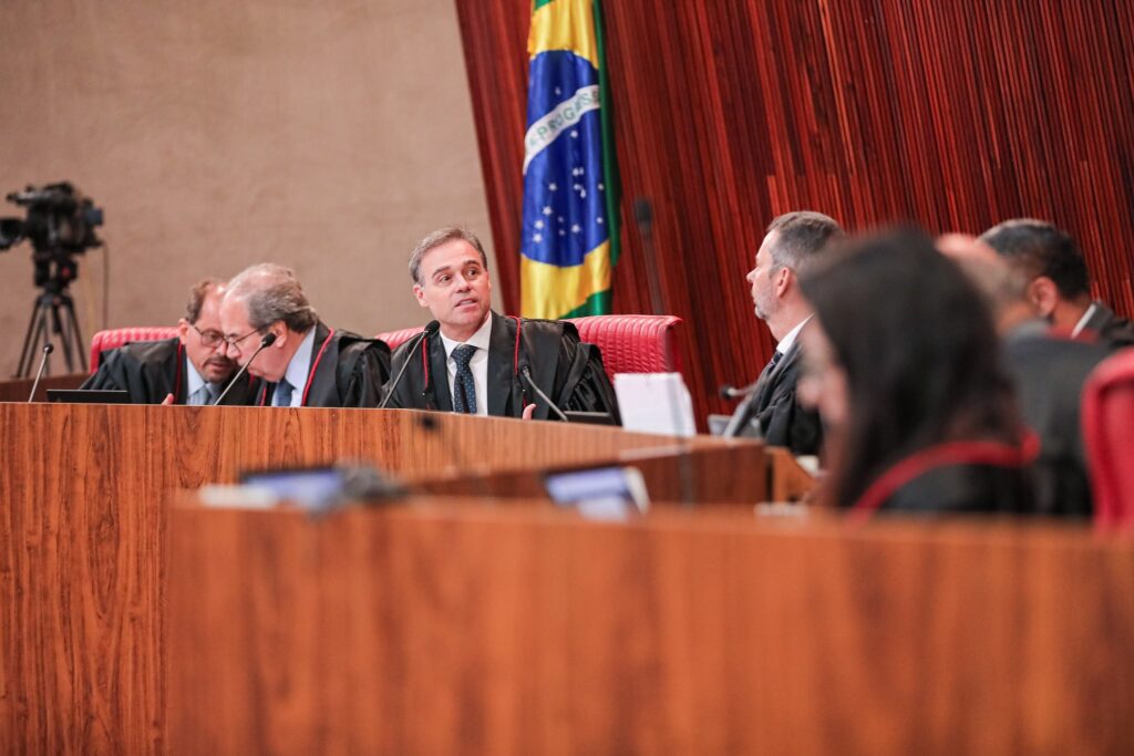 Sessão plenária da Justiça Eleitoral com ministros sentados à mesa principal e bandeira do Brasil.