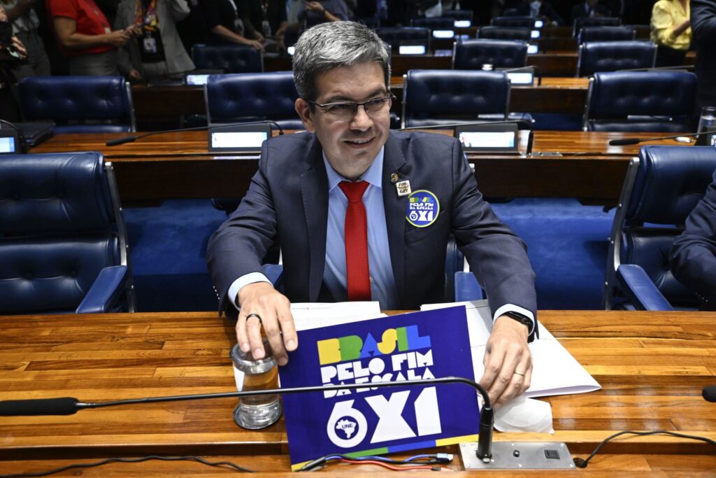 O senador Randolfe Rodrigues, vestindo terno e gravata vermelha, segura um cartaz com os dizeres "Brasil pelo fim da escala 6x1" em sua bancada no Plenário do Senado Federal.