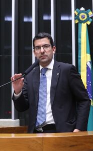 Deputado Marcelo Queiroz fala ao microfone em sessão deliberativa no Plenário da Câmara dos Deputados, com a bandeira do Brasil ao fundo.