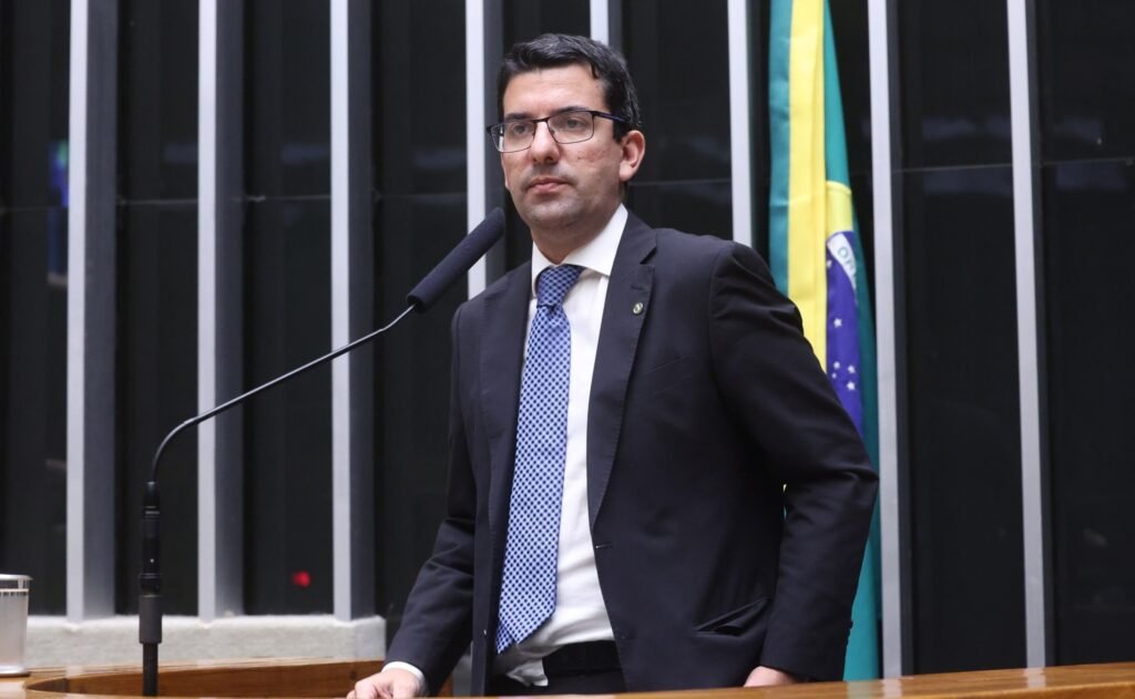 Marcelo Queiroz, de terno e óculos, fala ao microfone em plenário legislativo com a bandeira do Brasil ao fundo.