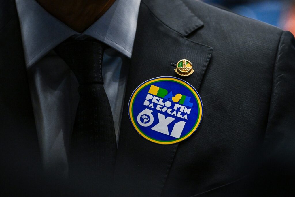 Pessoa anônima de terno e gravata exibe adesivo circular com os dizeres "Brasil pelo fim da escala 6x1" fixado na lapela.