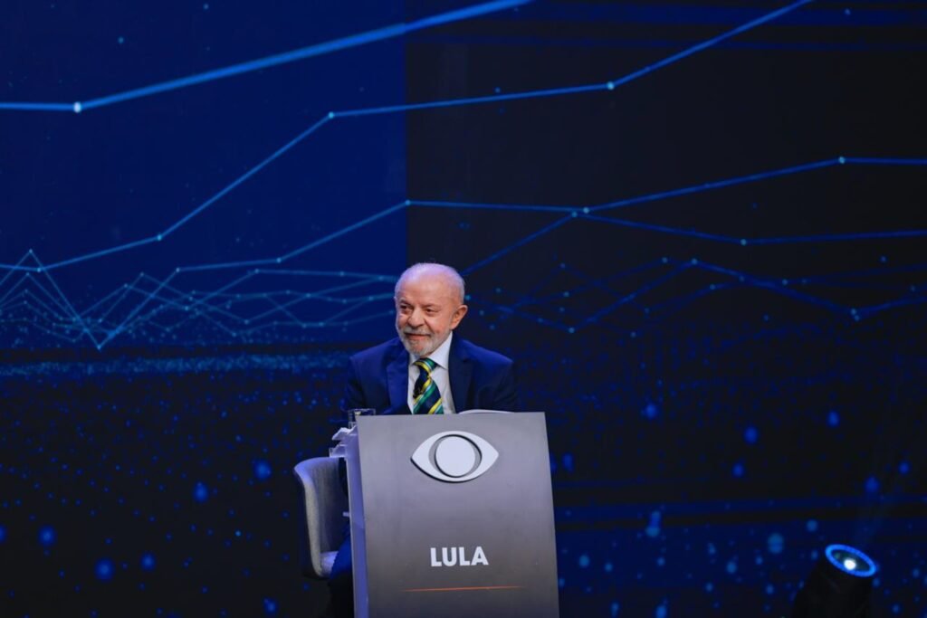 debate Lula e Flávio: O presidente Luiz Inácio Lula da Silva está sentado atrás de uma bancada com o logotipo da Band durante um debate televisivo.
